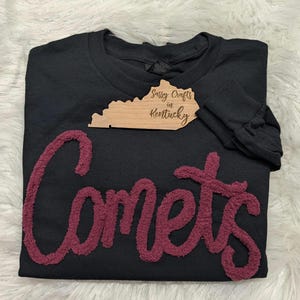 May include: A black long-sleeve sweatshirt featuring the word Comets in large, textured, maroon-coloured chenille lettering across the front. A wooden tag shaped like the state of Kentucky with the text Sassy Crafts in Kentucky is attached.
