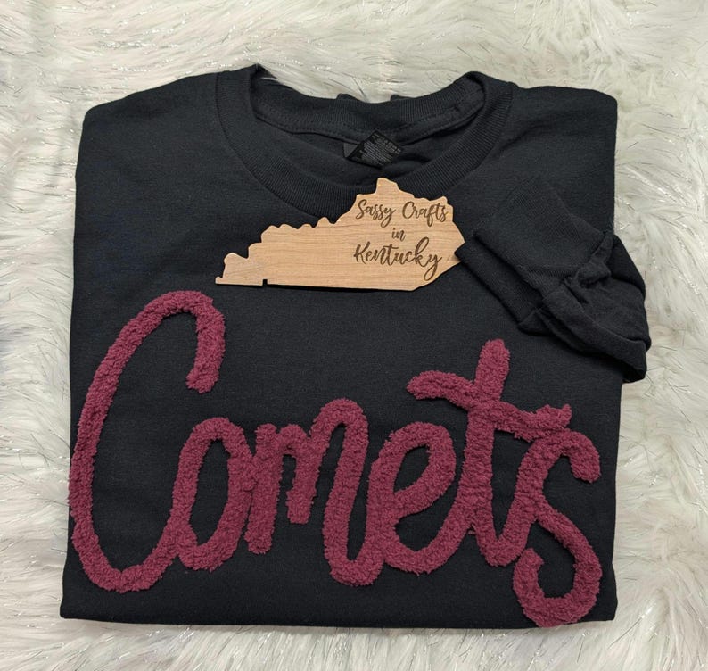 May include: A black long-sleeve sweatshirt featuring the word Comets in large, textured, maroon-coloured chenille lettering across the front. A wooden tag shaped like the state of Kentucky with the text Sassy Crafts in Kentucky is attached.