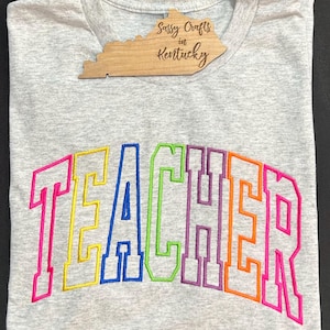May include: A gray t-shirt with the word "TEACHER" embroidered in rainbow colors. The letters are outlined in black and have a vintage, varsity-style font.