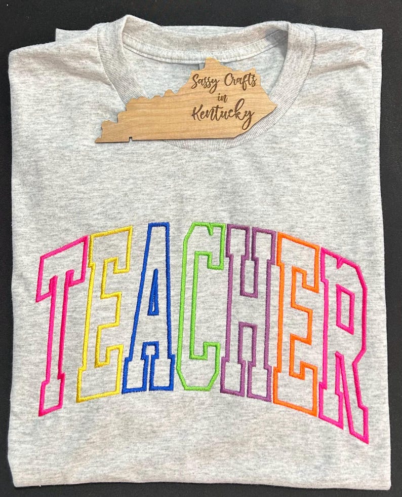 May include: A gray t-shirt with the word "TEACHER" embroidered in rainbow colors. The letters are outlined in black and have a vintage, varsity-style font.