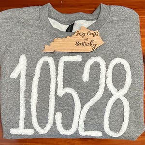 May include: A heather grey crewneck sweatshirt featuring the white fuzzy numbers 10528 across the front. A wooden cutout shaped like the state of Kentucky is placed near the neckline, with the text Sassy Crafts in Kentucky engraved on it.