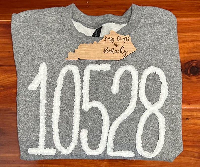 May include: A heather grey crewneck sweatshirt featuring the white fuzzy numbers 10528 across the front. A wooden cutout shaped like the state of Kentucky is placed near the neckline, with the text Sassy Crafts in Kentucky engraved on it.
