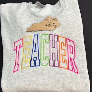 May include: A gray sweatshirt with the word "TEACHER" embroidered in rainbow colors. The word is in a large, arched font. The sweatshirt is from Sassy Crafts in Kentucky.