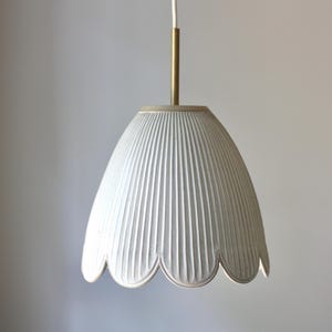 May include: A white, pendant light fixture with a scalloped edge and vertical ribbed texture. The light has a brass-colored stem and a white cord. The fixture is hanging against a plain, light-colored background.