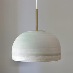 May include: A dome-shaped pendant light fixture with a matte, light gray ceramic shade featuring subtle horizontal ridges. The shade is suspended by a thin, vertical brass-colored metal rod that connects to a white electrical cord at the top.