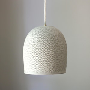 May include: A dome-shaped pendant light fixture featuring a matte off-white finish with a repeating embossed diamond pattern across the exterior surface. The light hangs from a single white cord attached to a small brass-colored metal fitting.