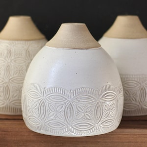 May include: Three white ceramic vases with a raised embossed pattern. The vases have a cream colored rim and are arranged on a wooden surface.
