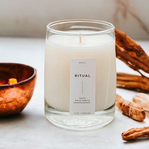 May include: A clear glass candle jar with a white label that reads "MOCC RITUAL SAGE PALO SANTO SANDALWOOD 12 OZ | COCO SOY". The candle is lit and the jar is sitting on a white surface with brown sticks.