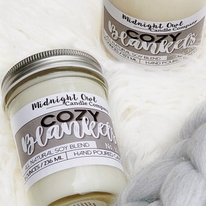 May include: Two glass candle jars with white wax and a lit wick. The jars are labeled "Midnight Owl Candle Company Cozy Blankets" and have a natural soy blend. The jars are sitting on a white fluffy surface.