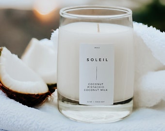 May include: A white candle in a clear glass jar with a white label that reads "SOLEIL COCONUT PISTACHIO COCONUT MILK 12 OZ COCO SOY". The candle is sitting on a white towel with a half coconut shell next to it.