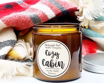 May include: A brown glass candle jar with a white label that reads "Midnight Owl Candle Company Cozy Cabin Soy Wax Candle 16 ounces". The candle is sitting on a plaid blanket.
