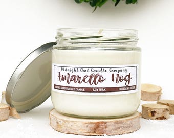 Peut inclure: Une bougie artisanale à la cire de soja de 14 onces dans un pot en verre transparent avec un couvercle argenté. La bougie est étiquetée "Midnight Owl Candle Company" et "Amaretto Nog" en texte brun. L'étiquette indique également "Holiday Edition".