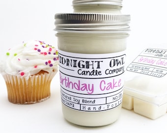 Könnte beinhalten: Eine weiße Kerze in einem Glasgefäß mit einem schwarz-weißen Etikett, auf dem "Midnight Owl Candle Company Birthday Cake" steht. Die Kerze ist eine natürliche Sojablendung und handgegossen. Das Gefäß steht auf einer weißen Oberfläche neben einem Cupcake mit weißem Frosting und bunten Streuseln.