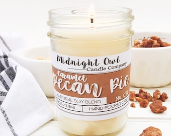 Puede incluir: Una vela en un tarro de vidrio con una etiqueta blanca que dice "Midnight Owl Candle Company" y "Caramel Pecan Pie". La vela está encendida y rodeada de trozos de nuez de pecán.