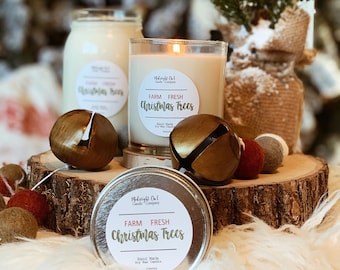 Puede incluir: Dos velas con etiquetas que dicen "Farm Fresh Christmas Trees" y una vela de hojalata con la misma etiqueta. Las velas están sobre una rodaja de madera con campanas doradas y pompones rojos y grises.