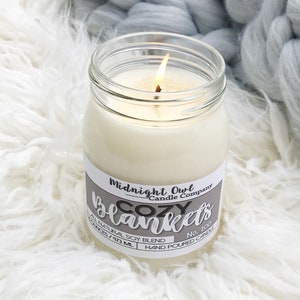 May include: A lit candle in a clear glass jar with a white label that reads "Cozy Blankets" and "Midnight Owl Candle Company." The candle is set on a white, fluffy surface with a gray, chunky knit blanket in the background. The candle is 16 ounces / 473 mL.