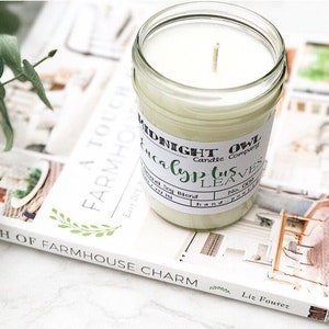 May include: A white candle in a clear glass jar with a white label that reads "Midnight Owl Candle Company Eucalyptus Leaves". The candle is sitting on a book titled "A Touch of Farmhouse Charm" by Liz Fourez.