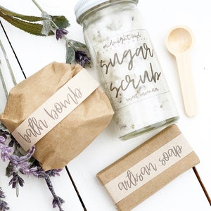 May include: A gift set of bath and body products wrapped in brown paper with labels. The set includes a bath bomb, a jar of sugar scrub labeled "midnight oat & sweet lavender sugar scrub", and a bar of artisan soap.