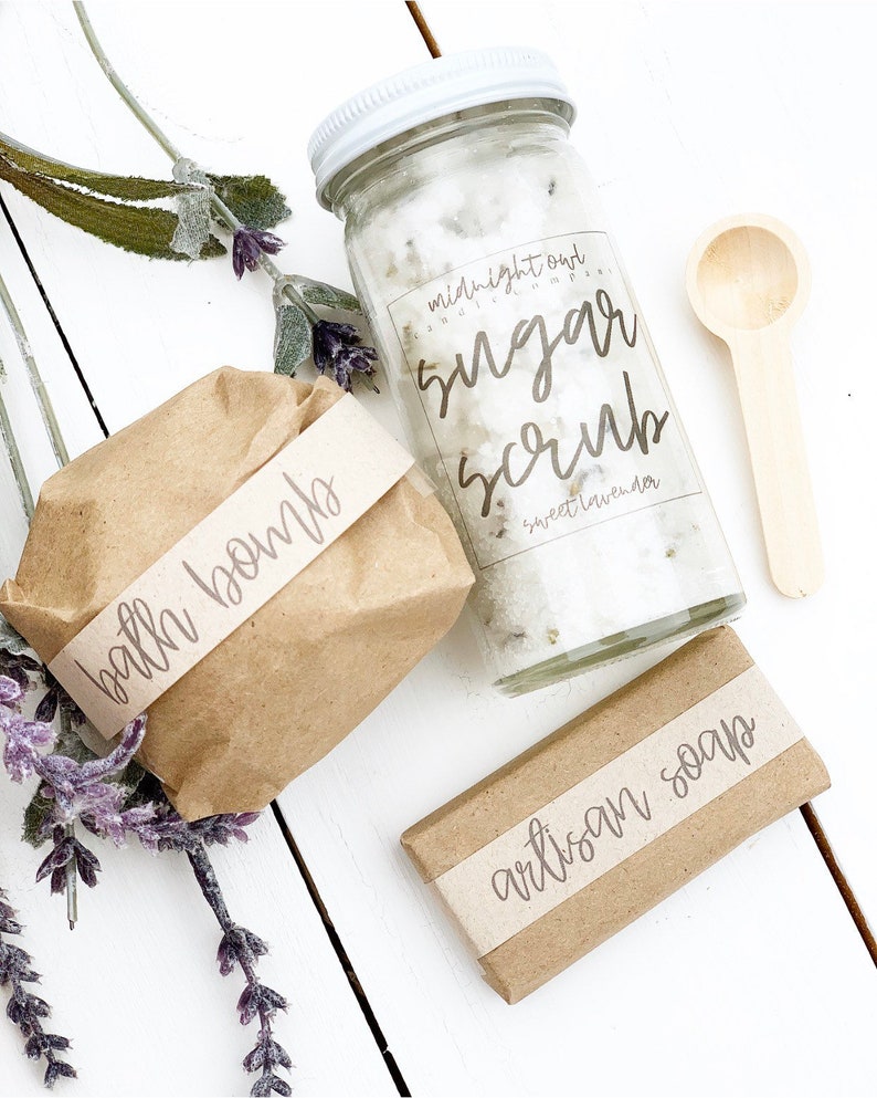 May include: A gift set of bath and body products wrapped in brown paper with labels. The set includes a bath bomb, a jar of sugar scrub labeled "midnight oat & sweet lavender sugar scrub", and a bar of artisan soap.