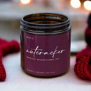 May include: A brown glass jar candle with a purple label that says "nutcracker" in white script. The label also says "Spiced Chai - Roasted Pumpkin - Oud" and "14 ounce | Coconut Soy Wax".