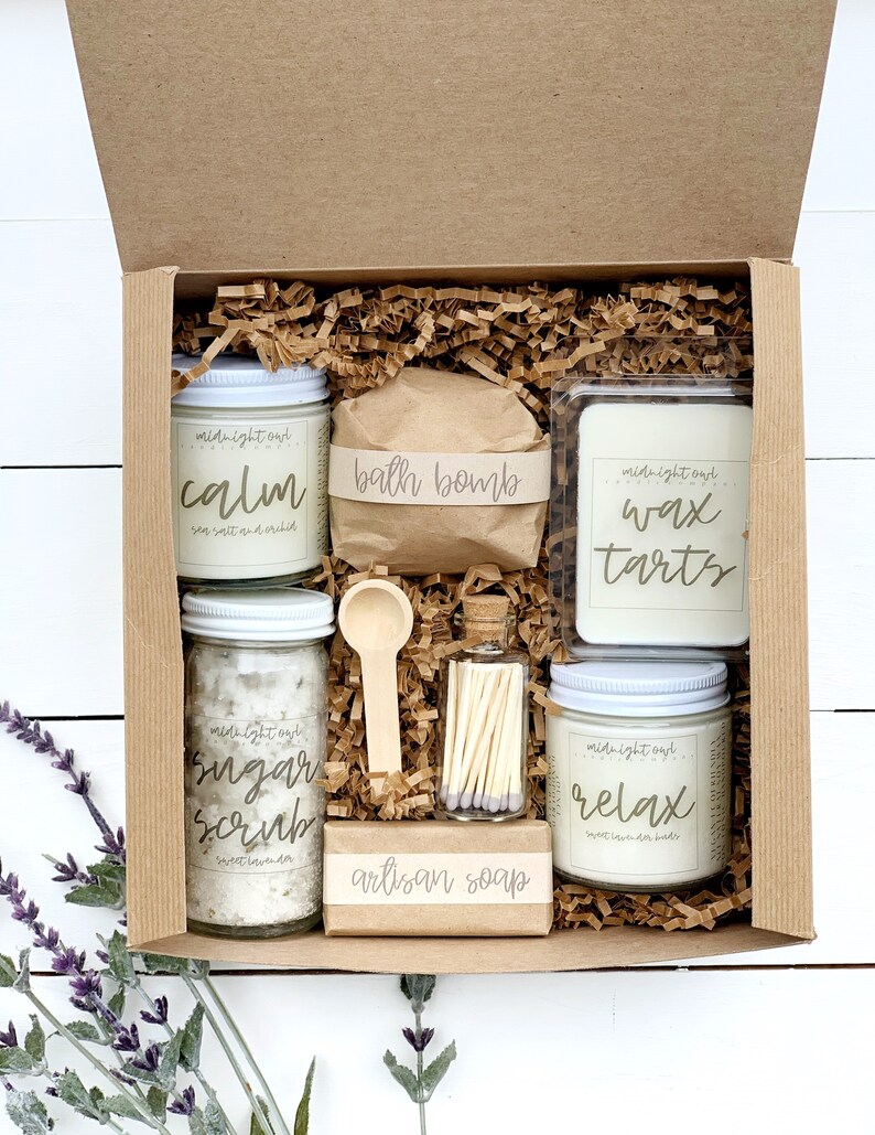 May include: A brown cardboard box filled with bath and body products. The box contains a bath bomb, a candle, a sugar scrub, a wax tart, a bar of soap, and a small jar of matches. The products are all labeled with the words "midnight owl" and have different scents, such as "calm", "relax", and "sugar scrub".