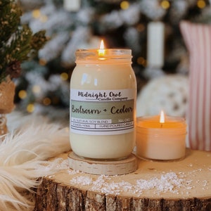 K&ouml;nnte beinhalten: Eine Kerze in einem durchsichtigen Glasgef&auml;&szlig; mit einem wei&szlig;en Etikett, auf dem "Midnight Owl Candle Company Balsam + Cedar No. 041 HAND POURED CANDLE" steht. Die Kerze ist angez&uuml;ndet und die Flamme ist sichtbar. Die Kerze steht auf einer Holzoberfl&auml;che mit wei&szlig;en Flocken.