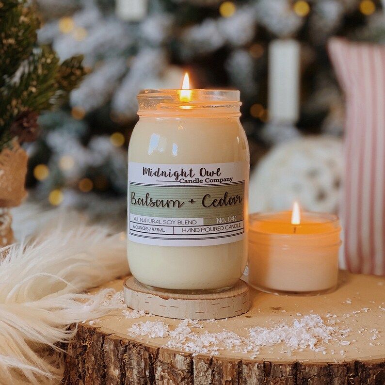K&ouml;nnte beinhalten: Eine Kerze in einem durchsichtigen Glasgef&auml;&szlig; mit einem wei&szlig;en Etikett, auf dem "Midnight Owl Candle Company Balsam + Cedar No. 041 HAND POURED CANDLE" steht. Die Kerze ist angez&uuml;ndet und die Flamme ist sichtbar. Die Kerze steht auf einer Holzoberfl&auml;che mit wei&szlig;en Flocken.