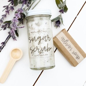 May include: A glass jar filled with white sugar scrub with a white lid and a label that reads "midnight owl candle company sugar scrub sweet lavender". A brown bar of soap with a label that reads "artisan soap" is next to the jar.