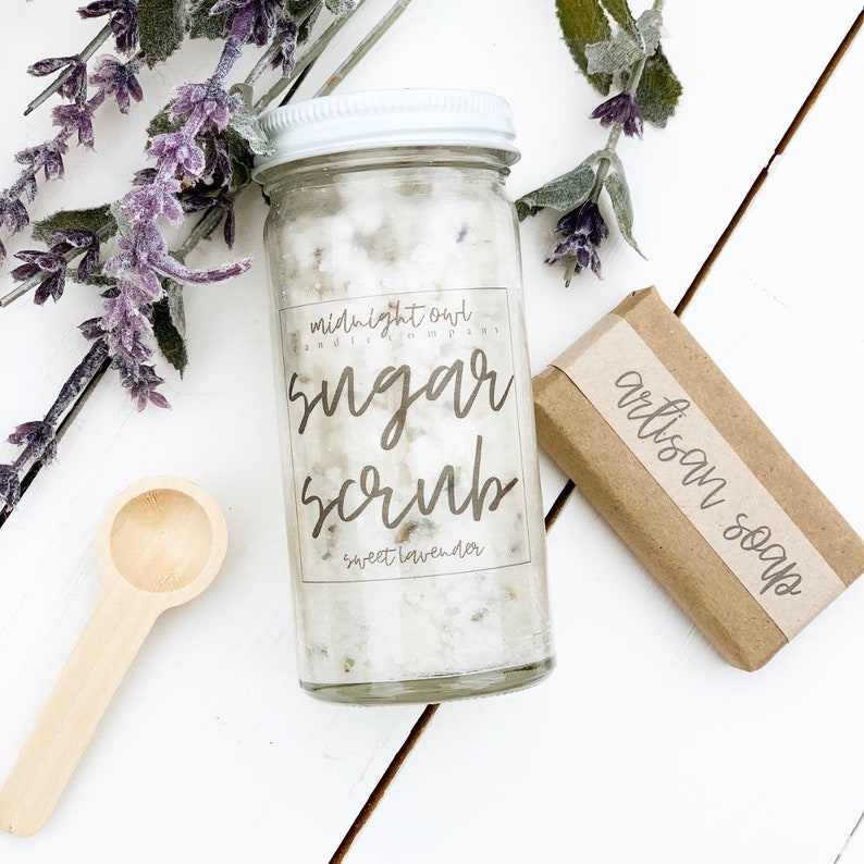 May include: A glass jar filled with white sugar scrub with a white lid and a label that reads "midnight owl candle company sugar scrub sweet lavender". A brown bar of soap with a label that reads "artisan soap" is next to the jar.