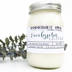 May include: A white candle in a clear glass jar with a silver lid. The label reads "MIDNIGHT OWL Candle Company Eucalyptus LEAVES All Natural Soy Blend No. 009 16 Ounces / 473 Ml hand-poured".