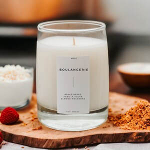 Puede incluir: Una vela blanca en un tarro de vidrio transparente con una etiqueta que dice "Mocc Boulangerie Pan horneado, Az&uacute;car de vainilla, Macarons de almendras 12 oz | Coco Soy". La vela est&aacute; sobre una superficie de madera con az&uacute;car moreno, un taz&oacute;n de copos de coco blancos y frambuesas rojas.