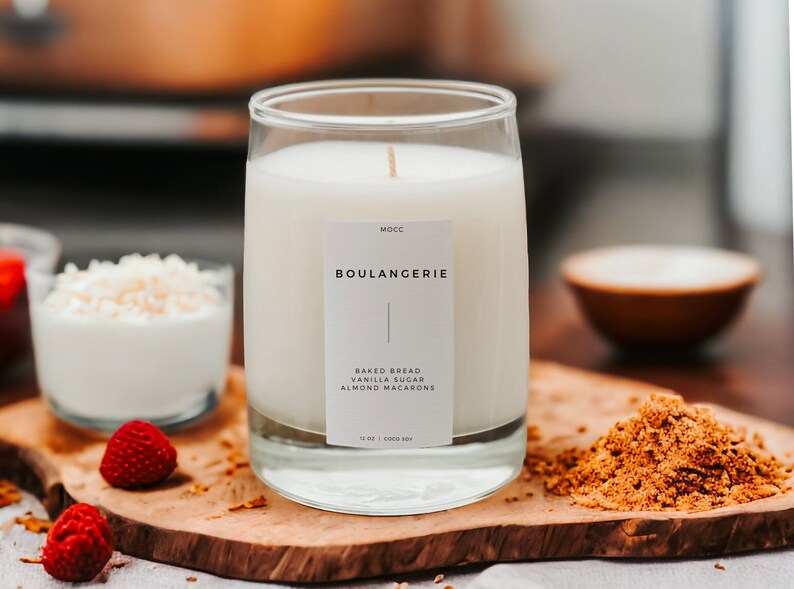 Puede incluir: Una vela blanca en un tarro de vidrio transparente con una etiqueta que dice "Mocc Boulangerie Pan horneado, Az&uacute;car de vainilla, Macarons de almendras 12 oz | Coco Soy". La vela est&aacute; sobre una superficie de madera con az&uacute;car moreno, un taz&oacute;n de copos de coco blancos y frambuesas rojas.
