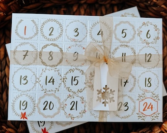 Könnte beinhalten: Ein weißer Adventskalender mit 24 nummerierten Türen, die mit grünen Kränzen und einem braunen Band verziert sind. Der Kalender ist mit einem braunen Band gebunden und hat ein Schneeflocken-förmiges Etikett.