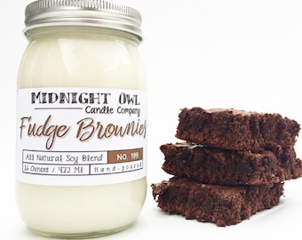 Könnte beinhalten: Eine handgegossene Sojawachskerze in einem durchsichtigen Glas mit silbernem Deckel. Die Kerze hat ein Fassungsvermögen von 473 ml. Das Etikett lautet "Midnight Owl Candle Company" und "Fudge Brownie" mit der Nummer "199". Im Hintergrund befinden sich drei Schokoladen-Brownies.