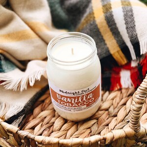 May include: A glass jar candle with a white label that reads "Midnight Owl Candle Company" and "Vanilla Tobacco No. 031". The label also says "Natural Soy Blend" and "8 oz / 237 ml - Hand Poured Candle". The candle is sitting on a woven basket.