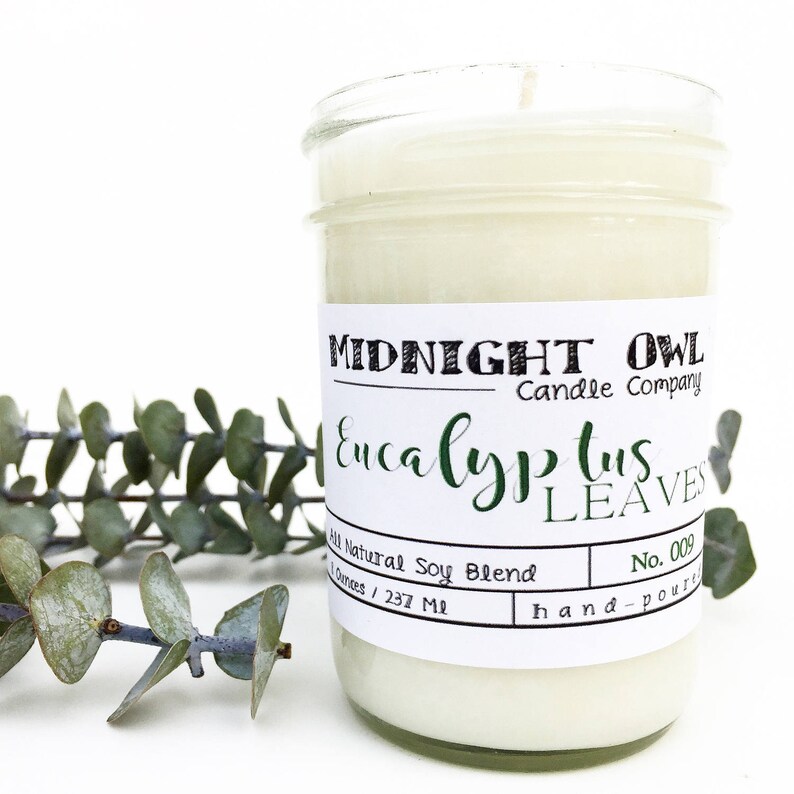 May include: A white candle in a clear glass jar with a white label that reads "MIDNIGHT OWL Candle Company Eucalyptus Leaves All Natural Soy Blend 8 ounces / 237 ml No. 009 hand-poured".