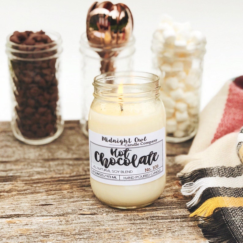 K&ouml;nnte beinhalten: Eine brennende Kerze in einem durchsichtigen Glasgef&auml;&szlig; mit einem wei&szlig;en Etikett, auf dem "Midnight Owl Candle Company Hot Chocolate No. 109 All Natural Soy Blend 8 Ounces / 213 ML Hand Poured Candle" steht.