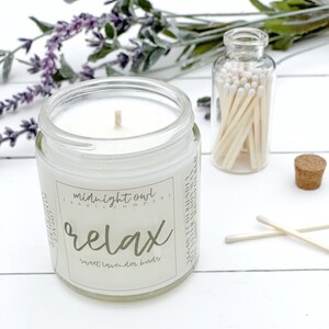 May include: A white candle in a glass jar with a label that reads "midnight owl candle company" and "relax sweet lavender buds". The candle is next to a small glass jar filled with matches.