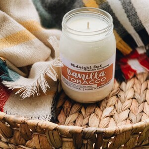 May include: A glass jar candle with a white label that reads "Midnight Owl Candle Company" and "Vanilla Tobacco No. 031". The candle is sitting on a woven basket with a blanket in the background.