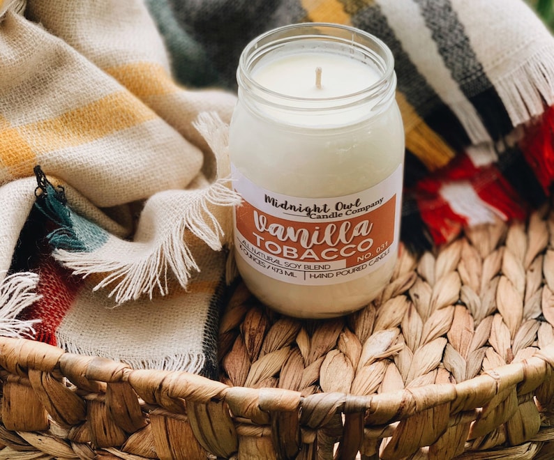 May include: A glass jar candle with a white label that reads "Midnight Owl Candle Company" and "Vanilla Tobacco No. 031". The candle is sitting on a woven basket with a blanket in the background.