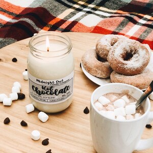 K&ouml;nnte beinhalten: Ein wei&szlig;es Kerzengef&auml;&szlig; mit einem schwarzen Etikett, auf dem "Midnight Owl Candle Company" und "Hot Chocolate" mit der Nummer "109" steht. Die Kerze ist angez&uuml;ndet und es gibt Marshmallows in einer Tasse hei&szlig;er Schokolade, Donuts mit Puderzucker und Schokoladenst&uuml;ckchen auf einer Holzoberfl&auml;che.