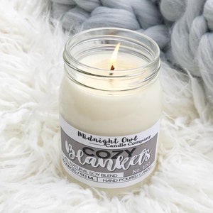 May include: A clear glass jar candle with a white label that reads "Midnight Owl Candle Company Cozy Blankets No. 100 All Natural Soy Blend 9.73 oz/273 ml Hand Poured Candle". The candle is lit and the flame is visible.