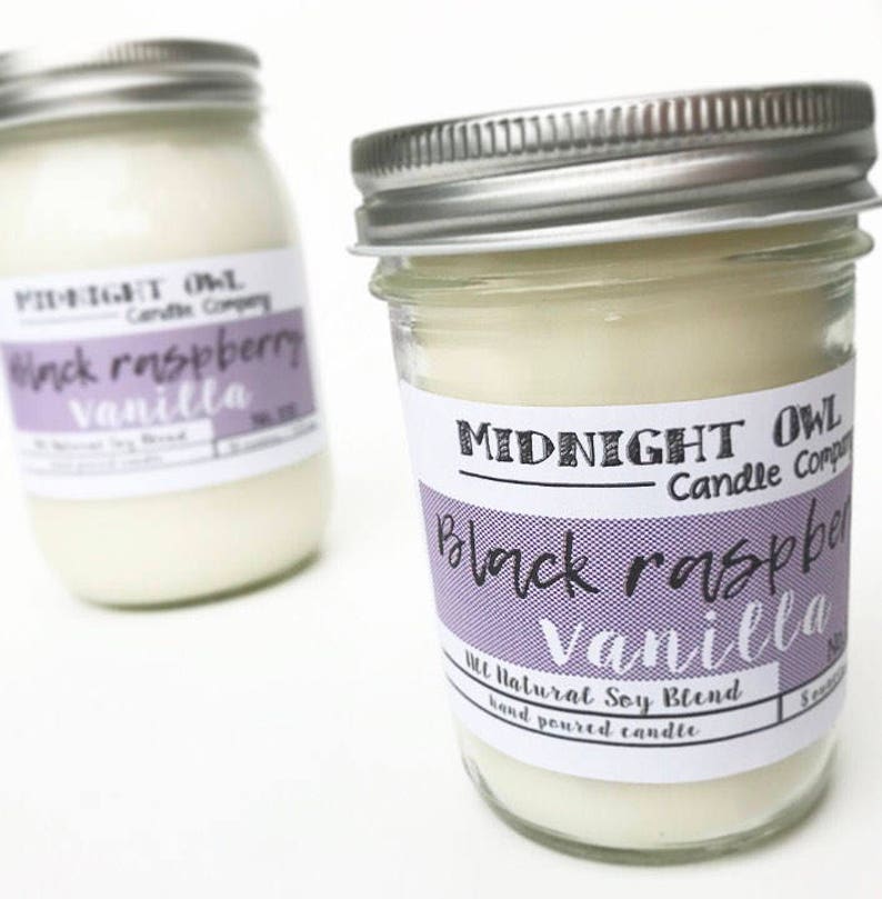 May include: Two glass jars with silver lids filled with a white candle. The labels on the jars read "Midnight Owl Candle Company" and "Black Raspberry Vanilla". The jars are on a white surface.