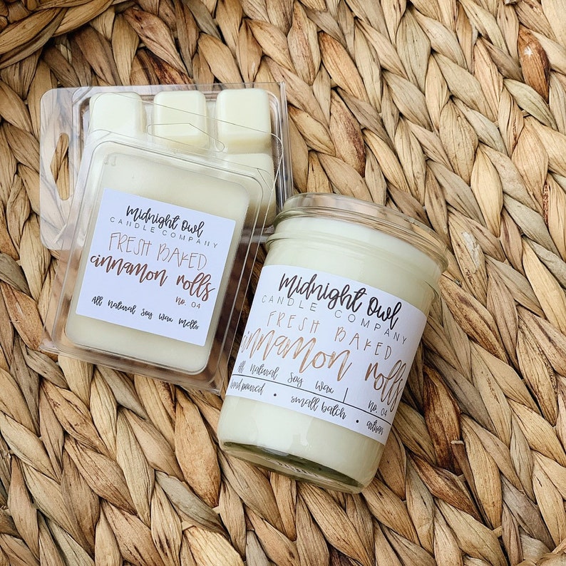 Puede incluir: Un juego de dos velas en frascos de vidrio con etiquetas que dicen "Midnight Owl Candle Company Fresh Baked Cinnamon Rolls No. 04". Las velas son de color amarillo claro y est&aacute;n sobre una superficie de mimbre tejido.