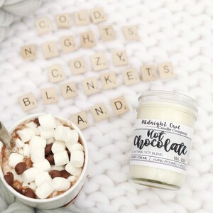 K&ouml;nnte beinhalten: Ein wei&szlig;es Kerzengef&auml;&szlig; mit einem schwarzen Etikett, auf dem "Midnight Owl Candle Company Hot Chocolate No. 109 All Natural Soy Blend Hand Poured Candle" steht. Die Kerze steht auf einer wei&szlig;en Strickdecke mit wei&szlig;en Buchstaben aus Keramik, die "Cold Nights Cozy Blankets And" buchstabieren. Eine Tasse hei&szlig;e Schokolade mit Marshmallows und Schokoladenst&uuml;ckchen steht ebenfalls auf der Decke.