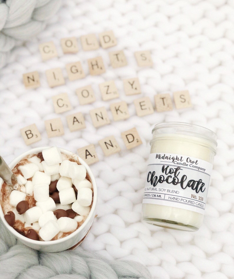 K&ouml;nnte beinhalten: Ein wei&szlig;es Kerzengef&auml;&szlig; mit einem schwarzen Etikett, auf dem "Midnight Owl Candle Company Hot Chocolate No. 109 All Natural Soy Blend Hand Poured Candle" steht. Die Kerze steht auf einer wei&szlig;en Strickdecke mit wei&szlig;en Buchstaben aus Keramik, die "Cold Nights Cozy Blankets And" buchstabieren. Eine Tasse hei&szlig;e Schokolade mit Marshmallows und Schokoladenst&uuml;ckchen steht ebenfalls auf der Decke.