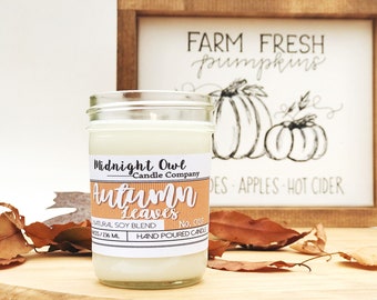 Puede incluir: Una vela en un tarro de vidrio con una etiqueta blanca que dice "Midnight Owl Candle Company Autumn Leaves No. 028 Natural Soy Blend Hand Poured Candle". La vela está sentada sobre una superficie de madera con hojas de otoño esparcidas a su alrededor. Un letrero de madera con el texto "FARM FRESH pumpkins DES APPLES HOT CIDER" está en el fondo.