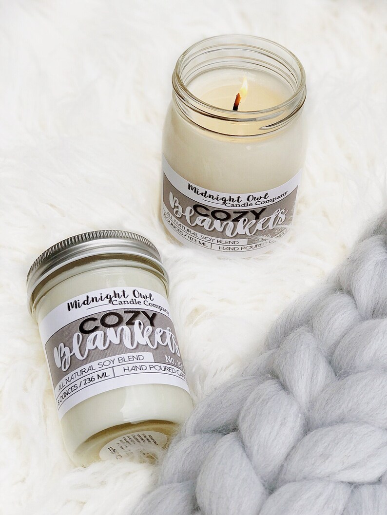 May include: Two glass candle jars with white wax and a lit wick. The jars are labeled "Midnight Owl Candle Company Cozy Blankets" and have a natural soy blend. The jars are sitting on a white fluffy surface.