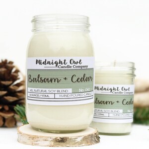 K&ouml;nnte beinhalten: Zwei Glasbeh&auml;lter mit wei&szlig;en Kerzen darin. Die Beh&auml;lter haben Etiketten mit der Aufschrift "Midnight Owl Candle Company" und "Balsam + Cedar". Die Etiketten sind gr&uuml;n und wei&szlig; mit einem Holzmaserungshintergrund. Die Beh&auml;lter stehen auf einer Holzoberfl&auml;che.