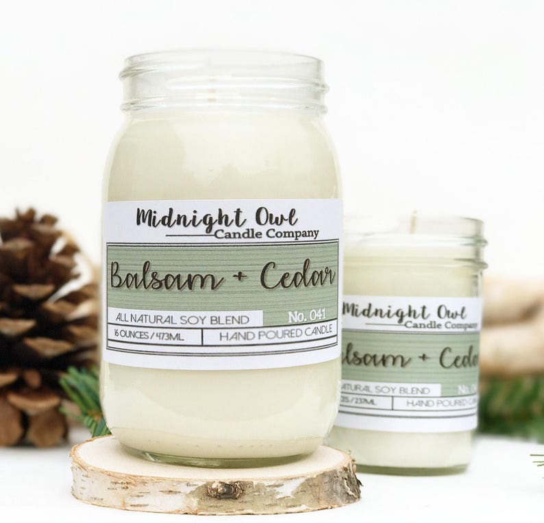 K&ouml;nnte beinhalten: Zwei Glasbeh&auml;lter mit wei&szlig;en Kerzen darin. Die Beh&auml;lter haben Etiketten mit der Aufschrift "Midnight Owl Candle Company" und "Balsam + Cedar". Die Etiketten sind gr&uuml;n und wei&szlig; mit einem Holzmaserungshintergrund. Die Beh&auml;lter stehen auf einer Holzoberfl&auml;che.